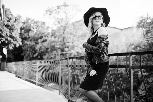 Blonde woman on black dress leather jacket sunglasses necklaces and hat against railings