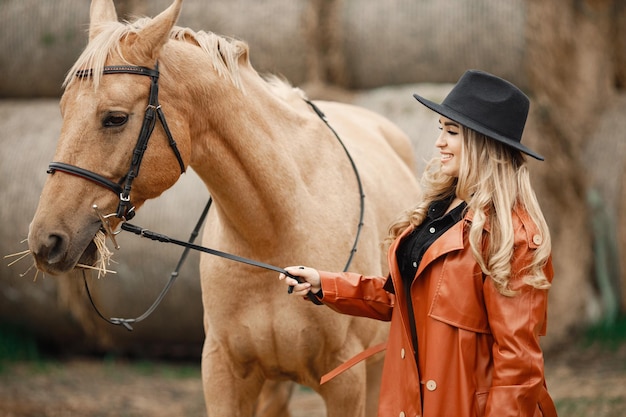 Free photo blonde woman and brown horse standing on a farm near hay bales. woman wearing black dress, red leather coat and hat. woman touching the horse.