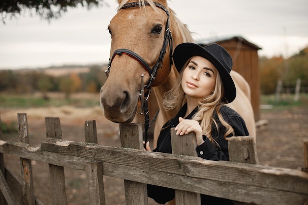 Blonde woman and brown horse standing on a farm. Woman wearing black clothes and hat. Woman touching the horse behinde the fence.