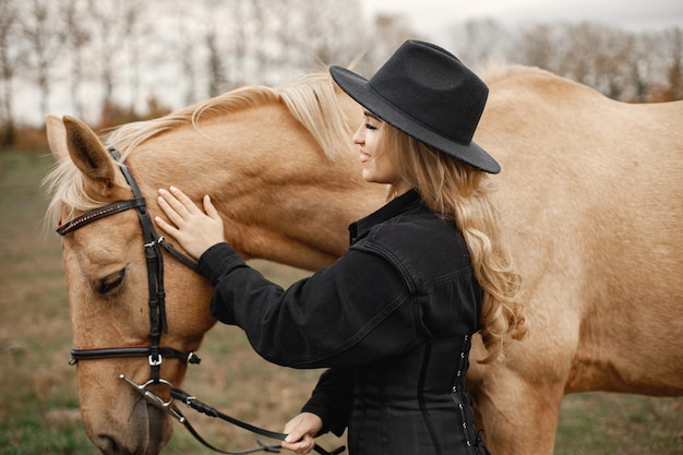 Blonde woman and brown horse standing in the field. Woman wearing black clothes and hat. Woman touching the horse.