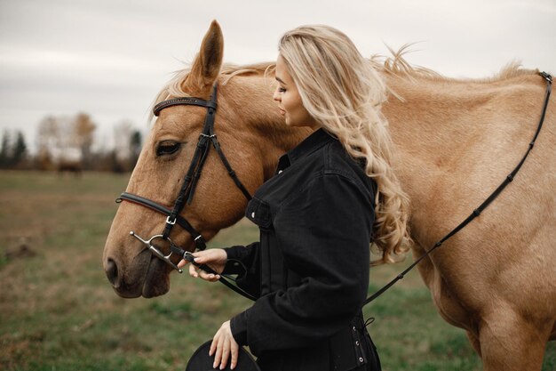 Blonde woman and brown horse standing in the field. Woman wearing black clothes. Woman touching the horse.