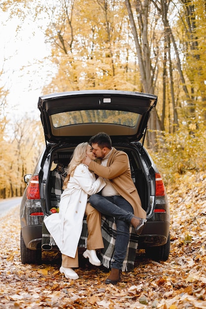 Free photo blonde woman and brunette man sitting in a black car trunk in autumn forest. woman wearing white coat and man beige coat. lovely couple kissing.