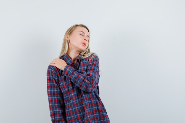 Blonde woman in checked shirt holding hand on shoulder and looking exhausted , front view.