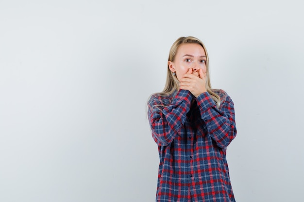 Blonde woman covering mouth with hands in checked shirt and looking surprised. front view.