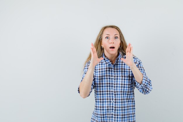 Blonde woman holding hands near face in shirt and looking surprised ,