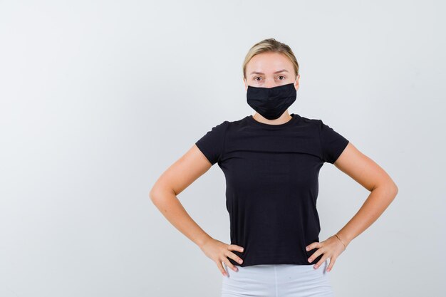 Blonde woman holding hands on waist in black t-shirt, white pants, black mask