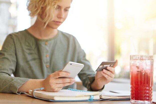 Blonde woman holding phone and credit card in cafe while having a drink