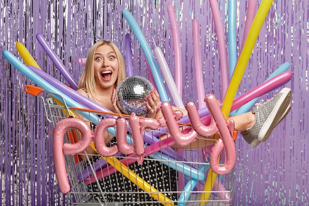 Free photo blonde woman at party in shopping cart holding balloons