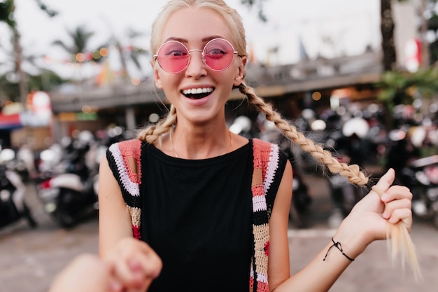 Free photo blonde woman in pink sunglasses posing with surprised smile. laughing woman with braids expressing amazement on blur street background.