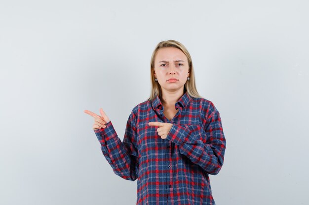 Blonde woman pointing left with index fingers and grimacing in checked shirt and looking displeased. front view.