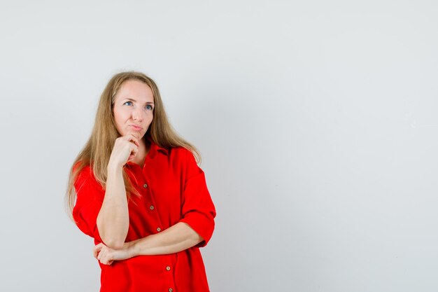 Blonde woman propping chin on hand in red shirt and looking hesitant ,