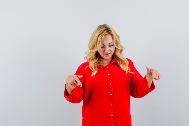 Blonde woman in red blouse pointing down with index fingers and looking happy