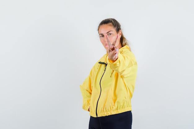 Blonde woman showing peace gesture in yellow bomber jacket and black pants and looking serious