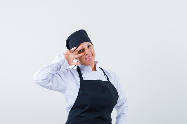 Blonde woman showing v sign on eye in black cook uniform and looking pretty.