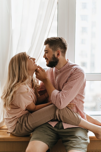 Free photo blonde woman sits on her boyfriend and laughs. man strokes with tenderness face of his beloved. portrait of couple against window.