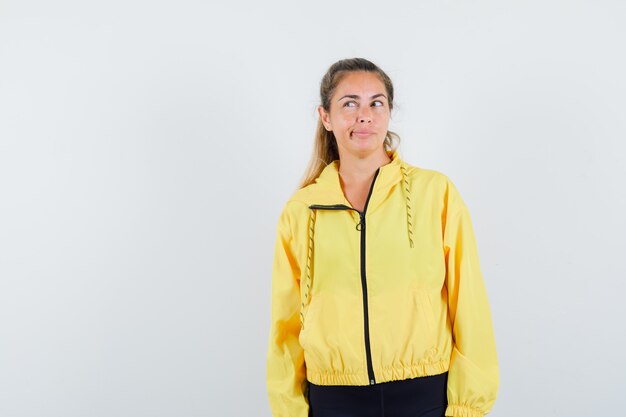 Blonde woman standing straight, looking away and posing at front in yellow bomber jacket and black pants and looking pensive