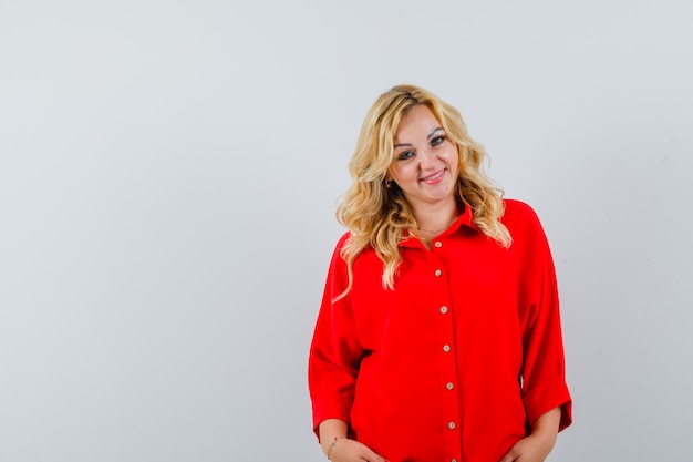 Blonde woman standing straight and posing at camera in red blouse and looking happy