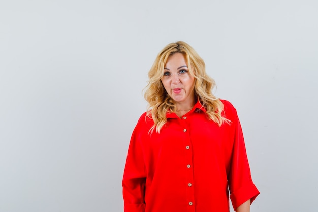 Blonde woman standing straight and posing at camera in red blouse and looking serious.