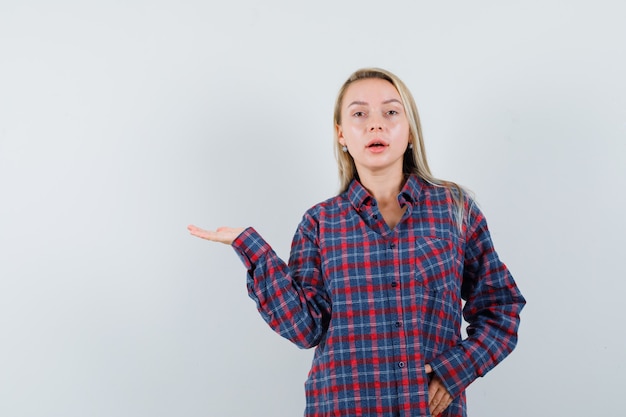 Blonde woman stretching hand as holding something and posing at camera in checked shirt and looking attractive. front view.