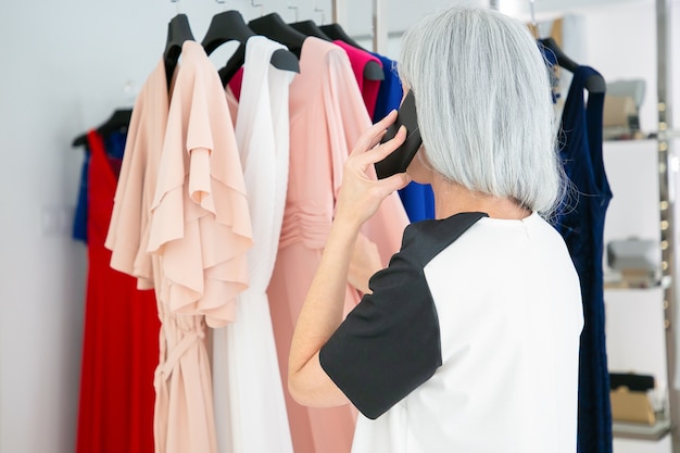 Blonde woman talking on mobile phone while choosing clothes and browsing dresses on rack in fashion store. Back view. Boutique customer or retail concept