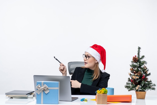 Blonde woman with a santa claus hat sitting at a table with a Christmas tree and a gift