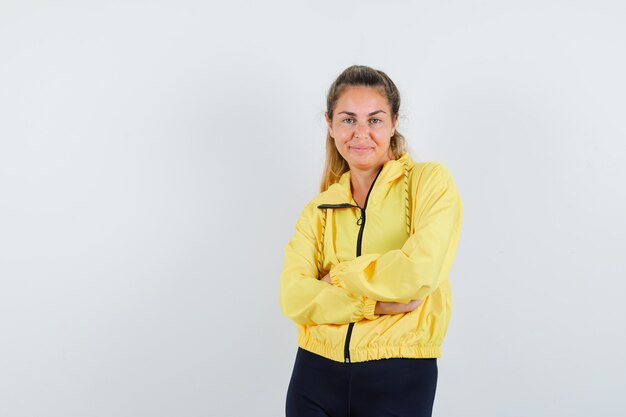 Blonde woman in yellow bomber jacket and black pants standing arms crossed and looking pretty