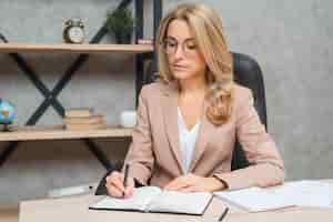 Free photo blonde young businesswoman writing on diary with pen at office