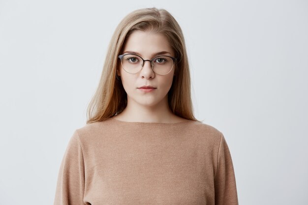 Blonde young female wearing stylish eyeglasses, with healthy skin looking directly . Serious confident girl with beautiful appearance dressed casually posing at studio. Beauty concept