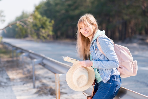 Free Photo blonde young woman holding hat and map standing near the road with her backpack looking at camera