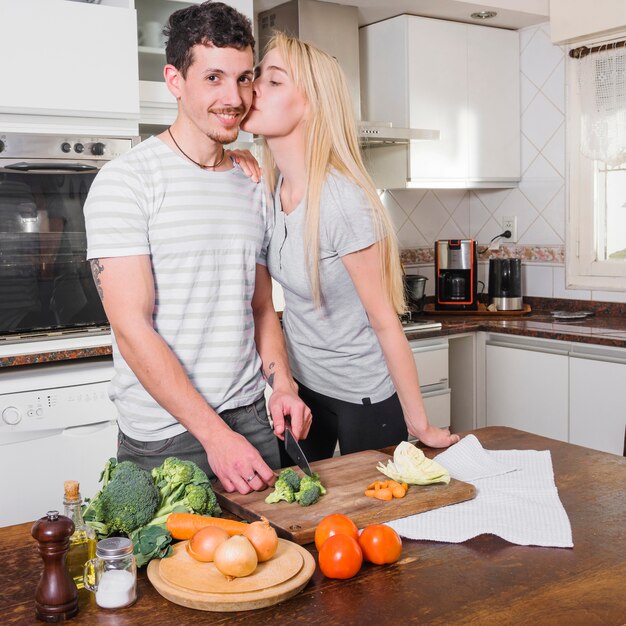 Blonde young woman kissing her husband cutting vegetable in the kitchen