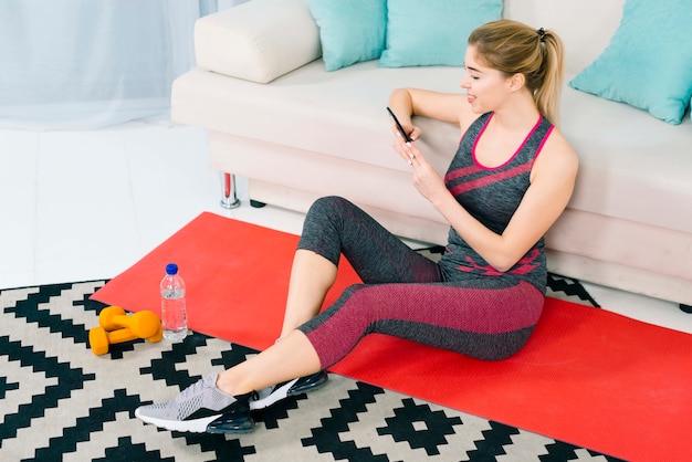 Blonde young woman sitting on carpet at home using mobile phone