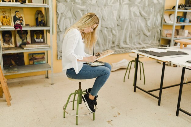 Blonde young woman sitting on stool painting sketch in workshop