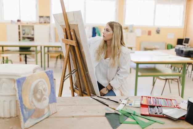 Blonde young woman sitting in workshop painting on easel