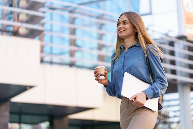 Blonde young woman smiling portrait holding laptop and coffee, wearing blue gentle shirt over modern building