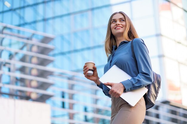 Blonde young woman smiling portrait holding laptop and coffee, wearing blue gentle shirt over modern building