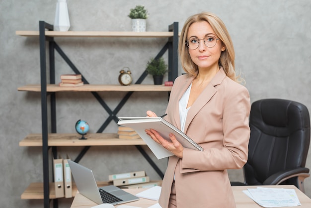 Free photo blonde young woman standing in front of office desk writing on spiral notebook