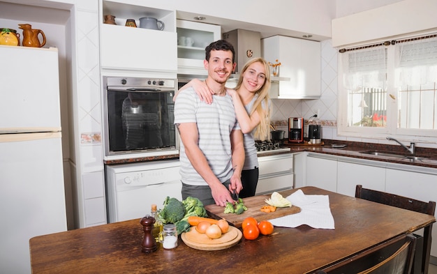 Free photo blonde young woman standing with her husband cutting vegetables in the kitchen