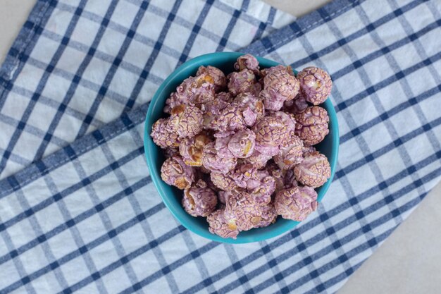 Blue bowl resting on a folded towel and filled with candy coated popcorn on marble table.