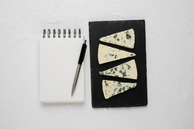 Free photo blue cheese slice arranged on black slate with empty spiral notebook and pen over concrete surface