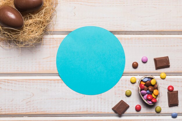 Blue circular frame with chocolates and easter eggs on nest over the wooden desk