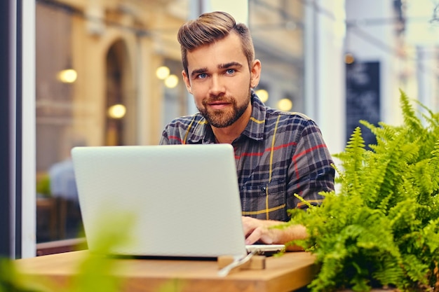 Free photo blue eyed, bearded male using a laptop in a cafe on a street.