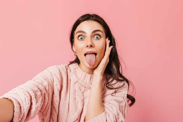 Blue-eyed emotional girl shows tongue. Woman in light sweater makes selfie on pink background.