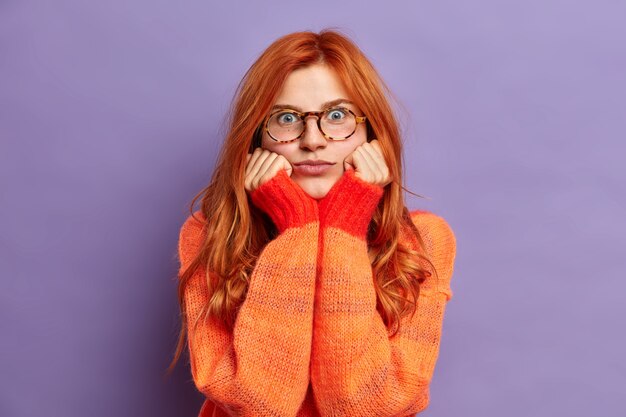 blue eyed pretty ginger girl keeps hands under chin and looks with great wonder focused dressed in warm sweater.