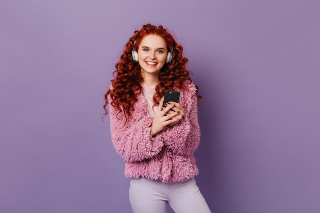 Blue-eyed sweet girl looks at camera on purple space with smile. Curly redhead woman in pink jacket posing in headphones with phone in her hands.