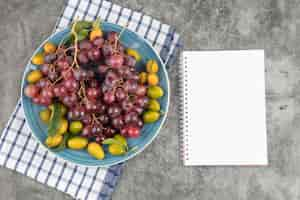 Free photo blue plate of kumquat fruits and red grapes with empty notebook.