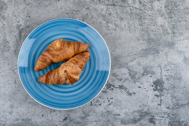 A blue plate of two plain fresh croissants on a stone table. 