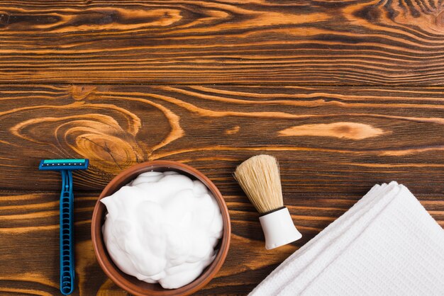 Blue razor; foam; shaving brush and white folded napkin against wooden surface