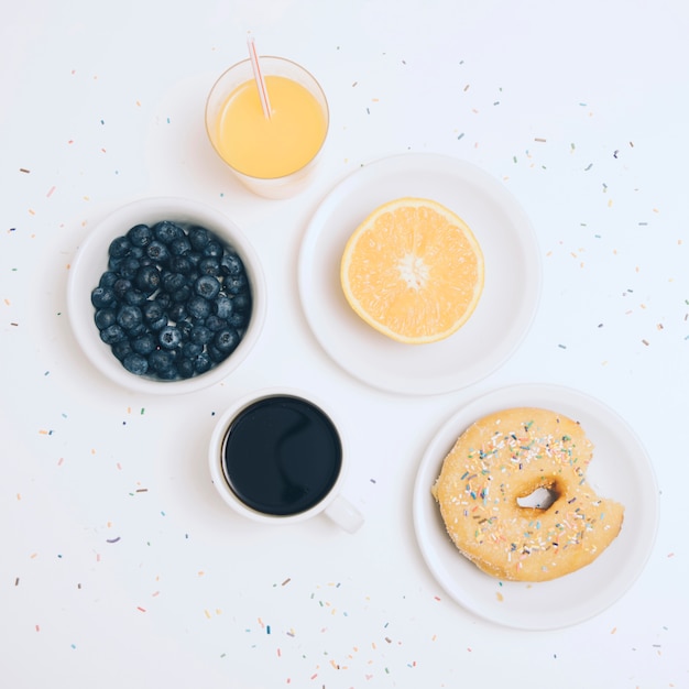 Free photo blueberries; coffee cup; halved orange; juice and donut with colorful sprinkles on white backdrop