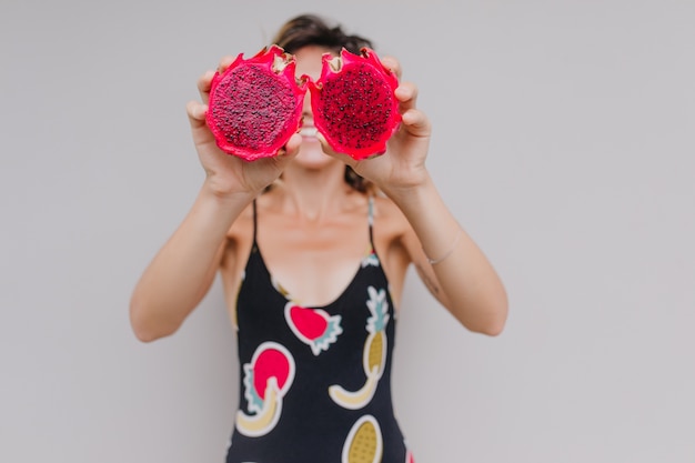 Free photo blur portrait of funny tanned woman with red pitaya in focus. indoor portrait of graceful girl in black dress holding dragon fruits.