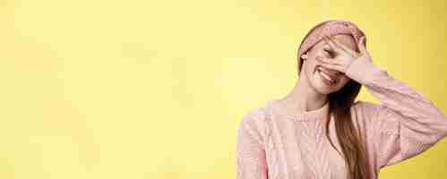 Free photo blushing cute girl feeling positive smiling happy joyful covering eyes with palm peeking through fingers delighted playful tilting head tender and lovely gazing at camera against yellow background